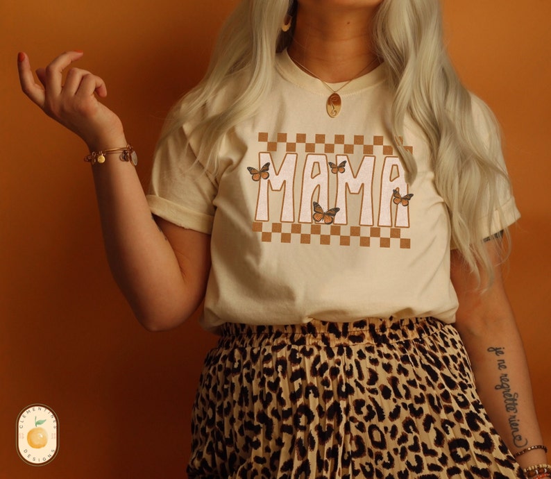 Puede incluir: Una camiseta de color crema con un patr&oacute;n a cuadros y la palabra "MAMA" en fuente marr&oacute;n. La camiseta tiene gr&aacute;ficos de mariposas. La modelo lleva una falda de estampado de leopardo.