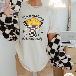 May include: Cream-colored t-shirt with the text "Keep making Lemonade" and a graphic of lemons in a checkered bowl. The person is wearing a brown and white checkered jacket and holding a black and white checkered phone case.