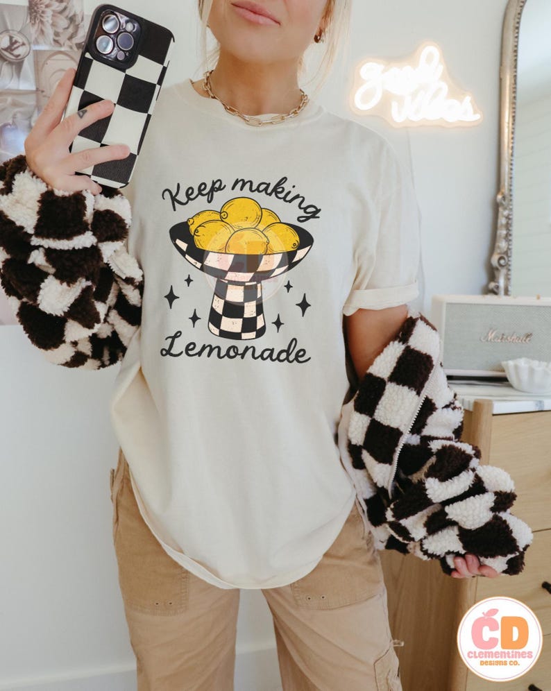 May include: Cream-colored t-shirt with the text "Keep making Lemonade" and a graphic of lemons in a checkered bowl. The person is wearing a brown and white checkered jacket and holding a black and white checkered phone case.