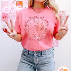 K&ouml;nnte beinhalten: Ein pinkfarbenes T-Shirt mit einem Motiv von Erdbeeren mit Schleifen. Die Erdbeeren sind in Schwarz umrandet und die Schleifen sind mit Schwarz gef&uuml;llt. Das Shirt wird von einer Person mit langen blonden Haaren und einer blauen Jeans getragen.