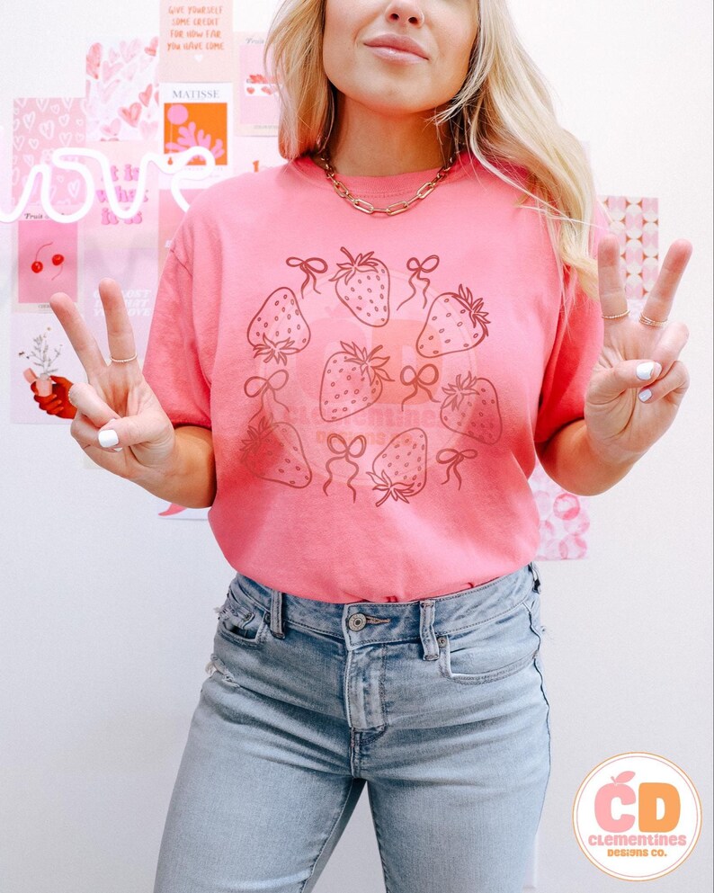 K&ouml;nnte beinhalten: Ein pinkfarbenes T-Shirt mit einem Motiv von Erdbeeren mit Schleifen. Die Erdbeeren sind in Schwarz umrandet und die Schleifen sind mit Schwarz gef&uuml;llt. Das Shirt wird von einer Person mit langen blonden Haaren und einer blauen Jeans getragen.