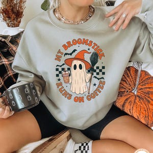 May include: A light grey sweatshirt with a Halloween design. The graphic shows a ghost in a witch's hat holding a broom and coffee. The text reads "MY BROOMSTICK RUNS ON COFFEE".