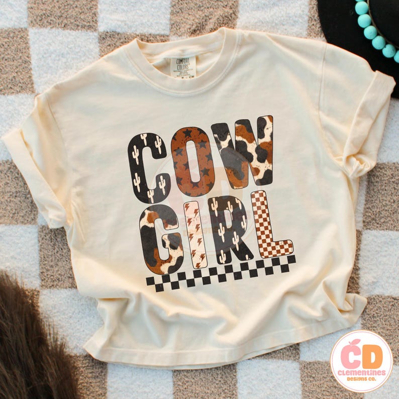 Puede incluir: Camiseta color crema con las palabras "COW GIRL" en un dise&ntilde;o audaz y multitexturizado. Las letras presentan patrones de cactus, piel de vaca y cuadros. La camiseta tiene mangas cortas y un corte holgado.
