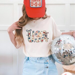 Pu&ograve; includere: Una maglietta bianca con una stampa floreale rosso, bianco e blu della parola "USA" sul davanti. La modella indossa un cappello da camionista rosso con il testo "Made in the U.S.A." sul davanti. La modella indossa anche una gonna di jeans blu chiaro.