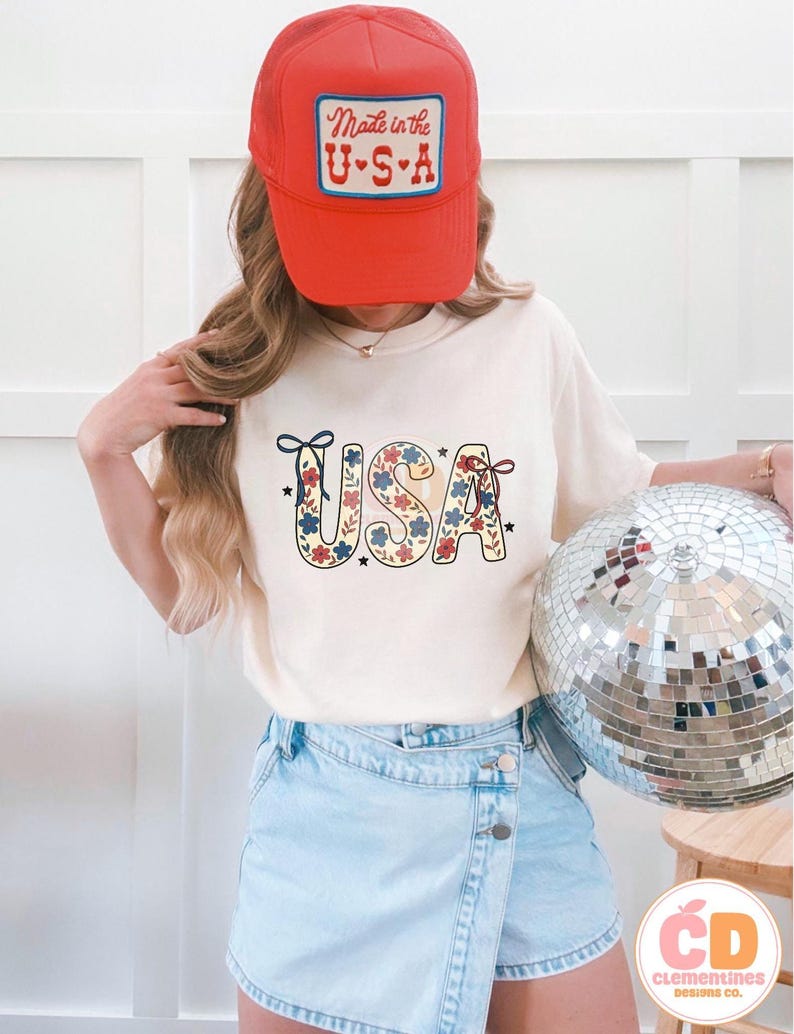 Pu&ograve; includere: Una maglietta bianca con una stampa floreale rosso, bianco e blu della parola "USA" sul davanti. La modella indossa un cappello da camionista rosso con il testo "Made in the U.S.A." sul davanti. La modella indossa anche una gonna di jeans blu chiaro.