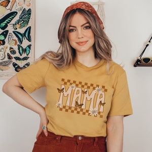 K&ouml;nnte beinhalten: Ein senfgelbes T-Shirt mit einem karierten Muster und dem Wort "MAMA" in einer Retro-Schrift. Das Design ist von Schmetterlingen und Blumen umgeben.