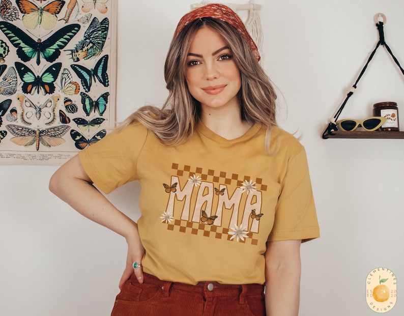 K&ouml;nnte beinhalten: Ein senfgelbes T-Shirt mit einem karierten Muster und dem Wort "MAMA" in einer Retro-Schrift. Das Design ist von Schmetterlingen und Blumen umgeben.