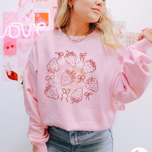 K&ouml;nnte beinhalten: Ein rosa Sweatshirt mit einem Aufdruck von Erdbeeren und Schleifen. Das Sweatshirt ist hellrosa und hat einen Rundhalsausschnitt. Die Erdbeeren sind dunkelrosa und haben einen wei&szlig;en Umriss. Die Schleifen sind hellrosa und haben einen wei&szlig;en Umriss.