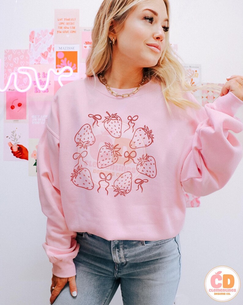 K&ouml;nnte beinhalten: Ein rosa Sweatshirt mit einem Aufdruck von Erdbeeren und Schleifen. Das Sweatshirt ist hellrosa und hat einen Rundhalsausschnitt. Die Erdbeeren sind dunkelrosa und haben einen wei&szlig;en Umriss. Die Schleifen sind hellrosa und haben einen wei&szlig;en Umriss.