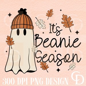 May include: A white ghost wearing an orange and brown beanie with the text "It's Beanie Season" in black lettering. The background is a light pink with brown and white leaves scattered around the ghost.