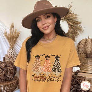May include: A woman wearing a mustard yellow t-shirt with a graphic of three ghosts wearing cowboy hats and the text "COWghoul". She is also wearing a brown fedora.
