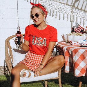 Pu&ograve; includere: Una donna indossa una maglietta rossa con una grafica floreale USA, una gonna a quadri rossi e bianchi e un bandana rosso. Tiene in mano una bottiglia di Coca-Cola ed &egrave; seduta su una sedia di vimini.