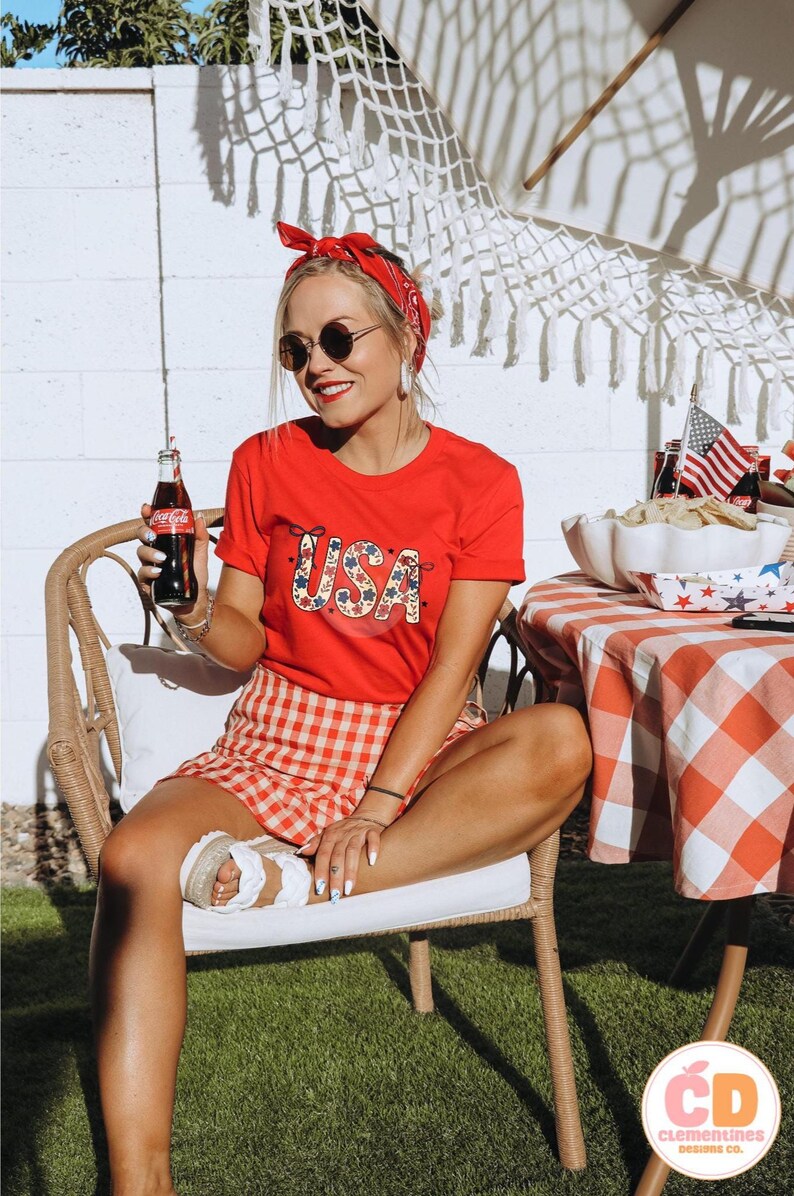 Pu&ograve; includere: Una donna indossa una maglietta rossa con una grafica floreale USA, una gonna a quadri rossi e bianchi e un bandana rosso. Tiene in mano una bottiglia di Coca-Cola ed &egrave; seduta su una sedia di vimini.