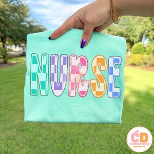 Pu&ograve; includere: Una maglietta verde acqua con la scritta "NURSE" in grandi lettere colorate a scacchi. Le lettere sono bordate di bianco e presentano sfumature di verde acqua, viola, rosa e arancione. La maglietta &egrave; tenuta da una mano con lo smalto viola.