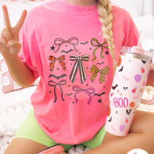 May include: A bright pink t-shirt featuring a collection of illustrated bows in various colors and patterns, with bats and stars. A pink and white tumbler with the word "BOO" and Halloween-themed graphics is held in the hand. The shirt is paired with neon green shorts.