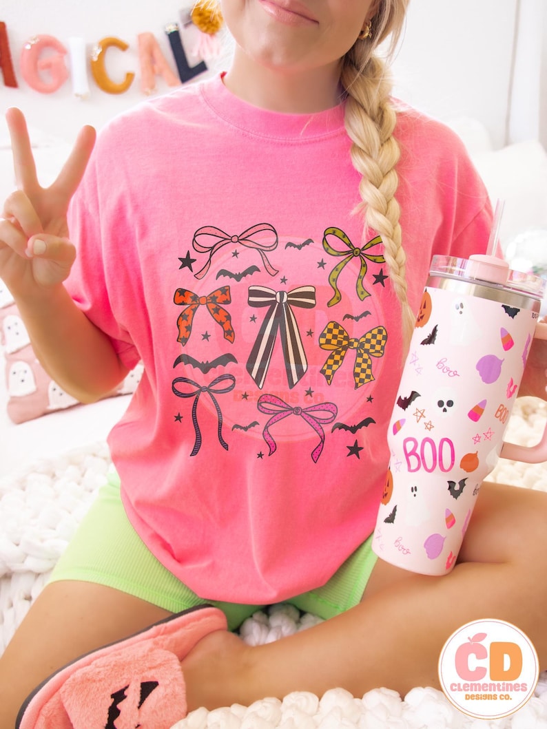 May include: A bright pink t-shirt featuring a collection of illustrated bows in various colors and patterns, with bats and stars. A pink and white tumbler with the word "BOO" and Halloween-themed graphics is held in the hand. The shirt is paired with neon green shorts.