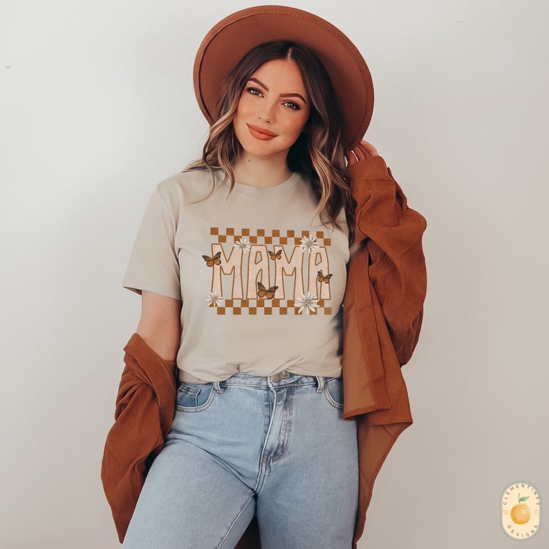 K&ouml;nnte beinhalten: Ein beiges T-Shirt mit einem karierten Muster und dem Wort "MAMA" in einer Retro-Schrift. Das Design zeigt braune Schmetterlinge und wei&szlig;e Blumen.