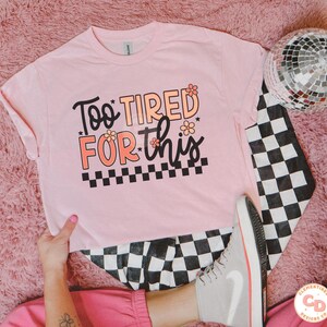 May include: Pink t-shirt with the text "Too Tired For This" in black with pink flowers and a black and white checkered pattern.