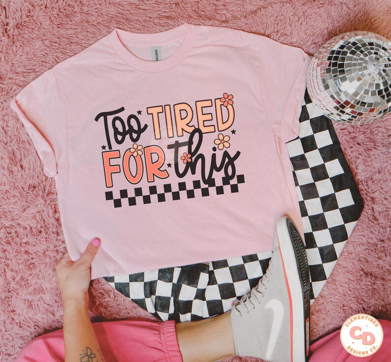 May include: Pink t-shirt with the text "Too Tired For This" in black with pink flowers and a black and white checkered pattern.