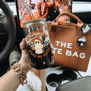 May include: A glass coffee cup with a bamboo lid and a glass straw. The cup features a Halloween design with a ghost, witch hat, and the text "My Broomstick Runs on Coffee". A brown tote bag and sunglasses are visible in the background.