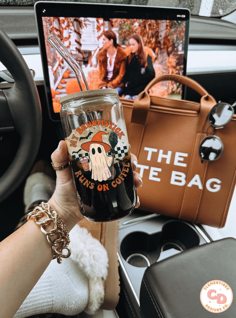 May include: A glass coffee cup with a bamboo lid and a glass straw. The cup features a Halloween design with a ghost, witch hat, and the text "My Broomstick Runs on Coffee". A brown tote bag and sunglasses are visible in the background.