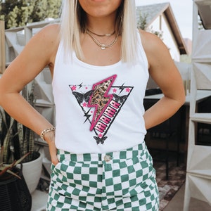 Pu&ograve; includere: Canotta bianca con una grafica di fulmine rosa e la parola "Mama" in lettere nere. La grafica presenta anche una stampa leopardata e farfalle.