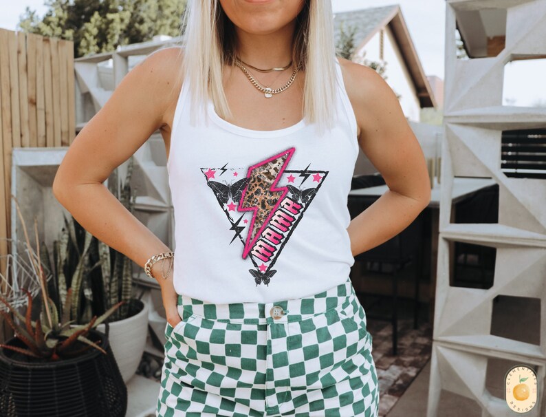 Pu&ograve; includere: Canotta bianca con una grafica di fulmine rosa e la parola "Mama" in lettere nere. La grafica presenta anche una stampa leopardata e farfalle.
