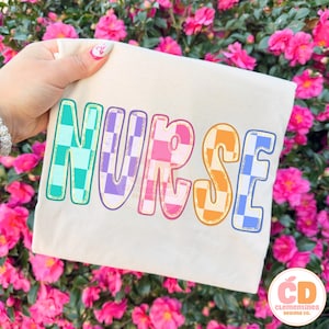 Pu&ograve; includere: Una maglietta color crema con la scritta "NURSE" in grandi lettere colorate a scacchi. Le lettere sono delineate in vari colori, tra cui verde acqua, viola, rosa, arancione e blu. La maglietta &egrave; tenuta contro uno sfondo di fiori rosa.