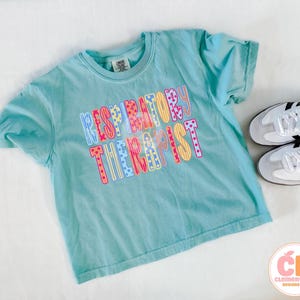 May include: A light blue t-shirt with the words "RESPIRATORY THERAPIST" in colorful, patterned letters. The shirt is paired with white sneakers with black stripes. The shirt is on a white surface.