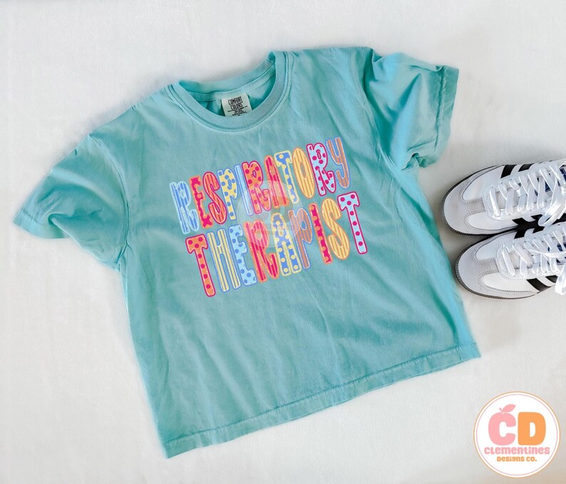May include: A light blue t-shirt with the words "RESPIRATORY THERAPIST" in colorful, patterned letters. The shirt is paired with white sneakers with black stripes. The shirt is on a white surface.