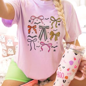 May include: A light purple t-shirt with a graphic of various bows, bats, and stars. A pink tumbler with the word "BOO" and Halloween-themed illustrations is held in the hand. The image also shows a pink slipper and a pillow with ghost designs.