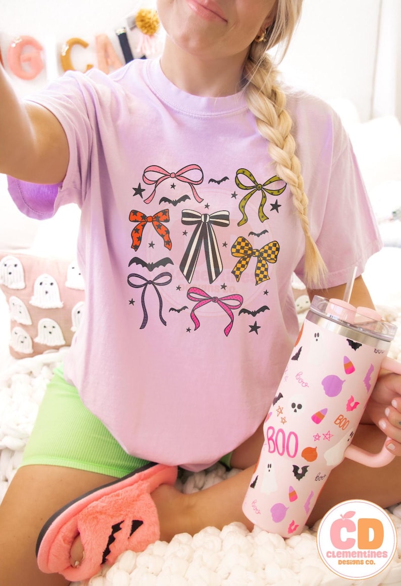 May include: A light purple t-shirt with a graphic of various bows, bats, and stars. A pink tumbler with the word "BOO" and Halloween-themed illustrations is held in the hand. The image also shows a pink slipper and a pillow with ghost designs.