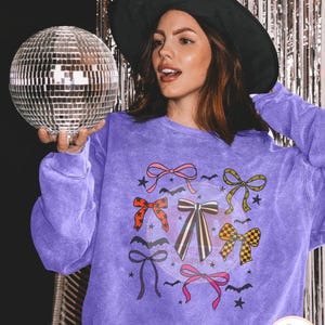 May include: A purple sweatshirt featuring a design of colorful bows, bats, and stars. The sweatshirt has a vintage wash effect. A woman wearing a black witch hat holds a disco ball.