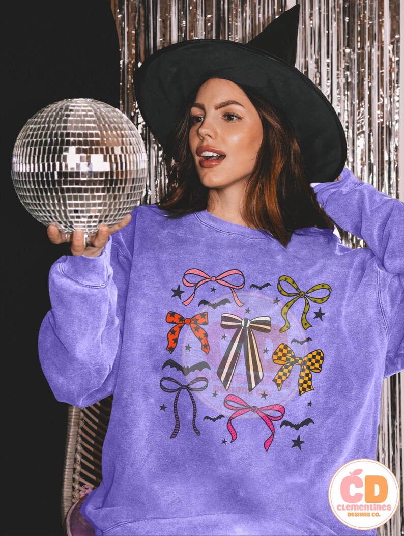 May include: A purple sweatshirt featuring a design of colorful bows, bats, and stars. The sweatshirt has a vintage wash effect. A woman wearing a black witch hat holds a disco ball.
