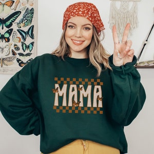 Puede incluir: Una sudadera verde oscuro con un dise&ntilde;o a cuadros marr&oacute;n y blanco y la palabra "MAMA" en una fuente retro. Mariposas con alas naranjas est&aacute;n dispersas alrededor del texto.