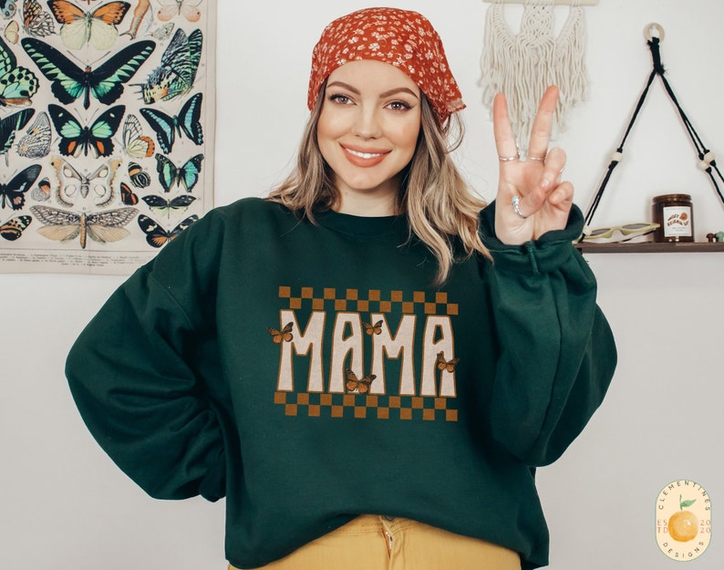 Puede incluir: Una sudadera verde oscuro con un dise&ntilde;o a cuadros marr&oacute;n y blanco y la palabra "MAMA" en una fuente retro. Mariposas con alas naranjas est&aacute;n dispersas alrededor del texto.