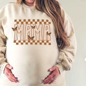 Puede incluir: Una sudadera beige con la palabra "MAMA" impresa en letras marrones con un patr&oacute;n a cuadros y mariposas marrones.