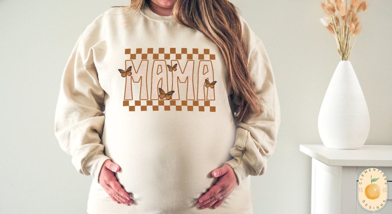 Puede incluir: Una sudadera beige con la palabra "MAMA" impresa en letras marrones con un patr&oacute;n a cuadros y mariposas marrones.