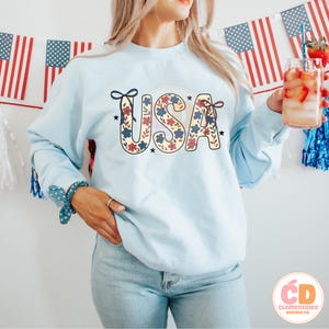 Pu&ograve; includere: Una felpa blu chiaro con un design floreale che compone la scritta "USA" in rosso, bianco e blu. La felpa &egrave; indossata da una persona con capelli lunghi castani e abbinata a jeans blu chiaro.