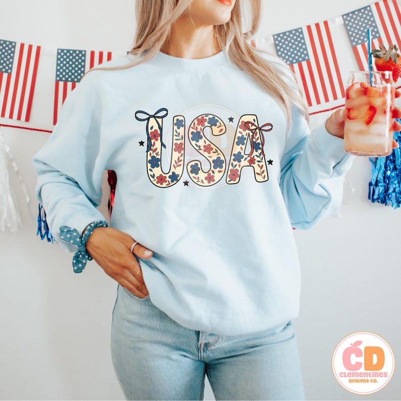 Pu&ograve; includere: Una felpa blu chiaro con un design floreale che compone la scritta "USA" in rosso, bianco e blu. La felpa &egrave; indossata da una persona con capelli lunghi castani e abbinata a jeans blu chiaro.
