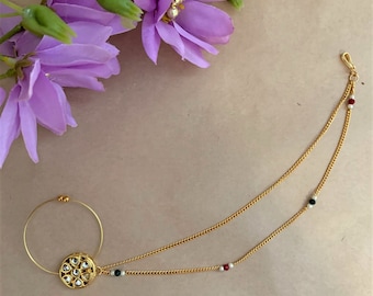 May include: A gold nose ring with a round, ornate design and a chain with small black, white, and red beads. The chain is attached to the nose ring and extends down to the shoulder.