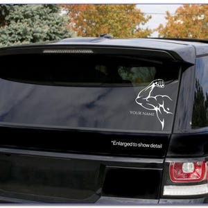 May include: White vinyl decal of a bicep flexing with the text "YOUR NAME" below. The decal is on the back window of a black car. The text "*Enlarged to show detail" is below the decal.