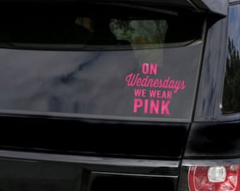 May include: A pink vinyl decal on the rear window of a black SUV. The decal reads "On Wednesdays We Wear Pink" in a cursive font. The vehicle's taillight is visible on the right side of the image.