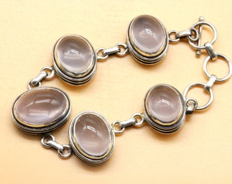 Puede incluir: Un brazalete de plata con cinco piedras de cuarzo rosa de forma ovalada. Cada piedra está engastada en un bisel de plata con un borde dorado. El brazalete tiene un cierre de palanca de plata.
