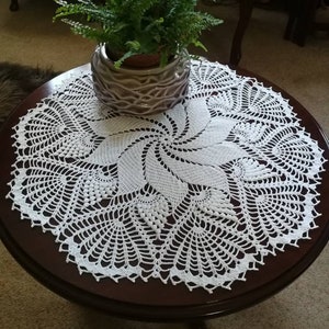 May include: White crocheted table runner with a spiral design. The runner is on a dark wood table with a potted fern plant in the center.