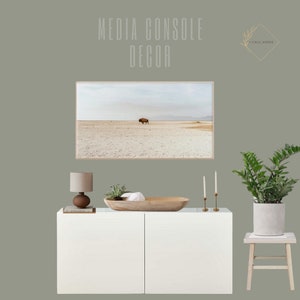 May include: White media console with two doors, a wooden bowl, a lamp with a beige shade, two candlesticks with white candles, and a potted plant with green leaves. The console is against a sage green wall with a framed photograph of a bison in a desert landscape above it. The text "MEDIA CONSOLE DECOR" is written in black above the photograph and the text "I CALL HOME" is written in black above the plant.
