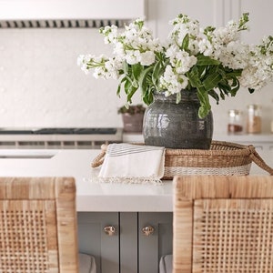 May include: White flowers in a gray ceramic vase on a woven tray with a white towel. The tray is on a white countertop with two woven chairs in the foreground.