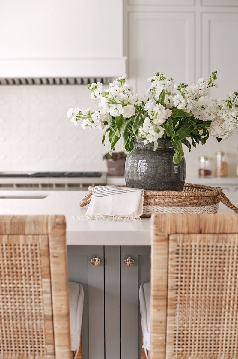 May include: White flowers in a gray ceramic vase on a woven tray with a white towel. The tray is on a white countertop with two woven chairs in the foreground.