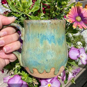 May include: A handcrafted ceramic mug with a turquoise and brown glaze. The mug features a distinctive, dripped design at the base and a robust handle. The mug is set amongst vibrant purple and pink flowers, creating a natural, artistic display.