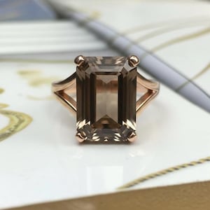 May include: A rose gold ring featuring a large, rectangular, smoky quartz gemstone. The gemstone is a translucent brown color and is set in a split-shank band. The ring has a classic, elegant design.